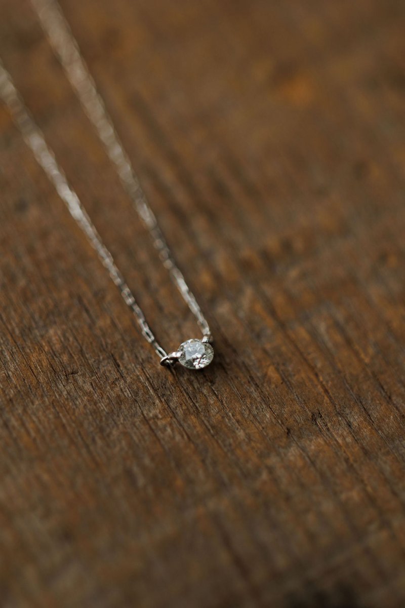 The Petite Diamond Necklace {White Gold} by Savannah Sevens western life{&}style, featuring a solitaire round diamond on an 18K white gold chain, rests elegantly on a rustic wooden surface.