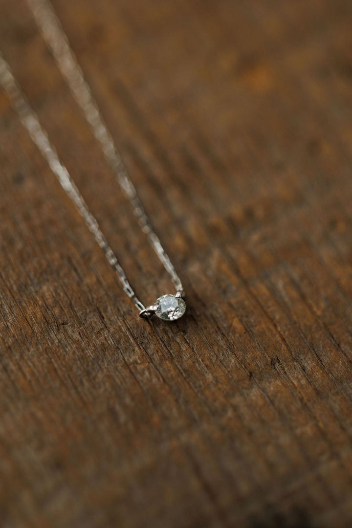 The Petite Diamond Necklace {White Gold} by Savannah Sevens western life{&}style, featuring a solitaire round diamond on an 18K white gold chain, rests elegantly on a rustic wooden surface.