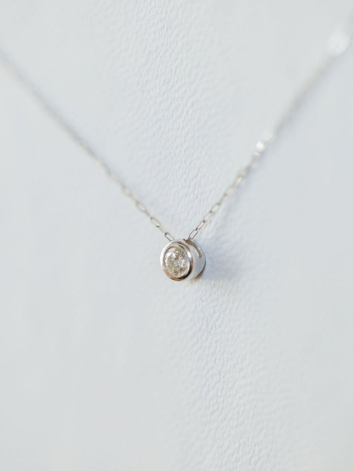 A close-up of the Savannah Sevens western life{&}style Petite Diamond Necklace {White Gold}, featuring a small round pendant with a natural round diamond, displayed against a light, textured background.