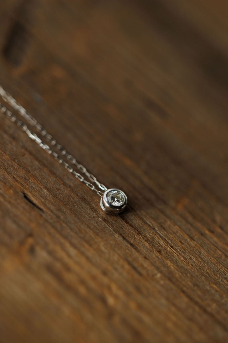 The Petite Diamond Necklace {White Gold} by Savannah Sevens western life{&}style features a natural round diamond in a small pendant, crafted from 18K white gold and displayed on a wooden surface.