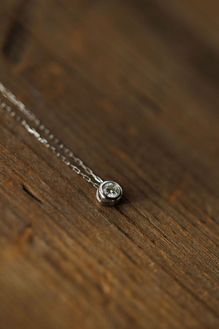 The Petite Diamond Necklace {White Gold} by Savannah Sevens western life{&}style features a natural round diamond in a small pendant, crafted from 18K white gold and displayed on a wooden surface.