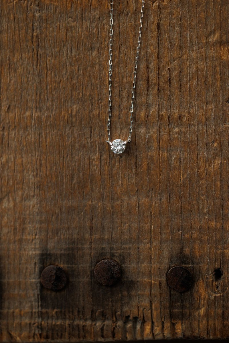 The Petite Diamond Necklace in White Gold by Savannah Sevens western life{&}style features a natural round diamond pendant, shining against a rustic wooden background.