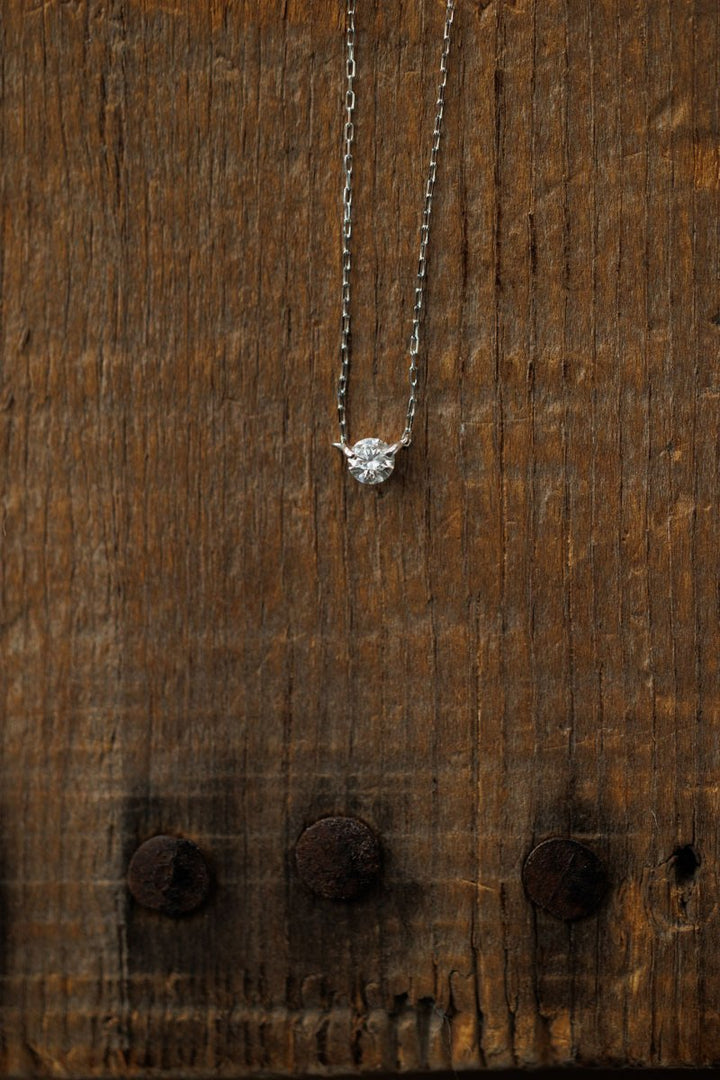 The Petite Diamond Necklace in White Gold by Savannah Sevens western life{&}style features a natural round diamond pendant, shining against a rustic wooden background.