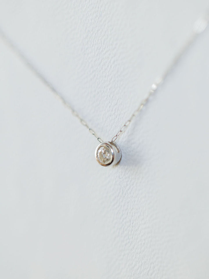A close-up of the Savannah Sevens western life{&}style Petite Diamond Necklace {White Gold}, featuring a small round pendant with a natural round diamond, displayed against a light, textured background.