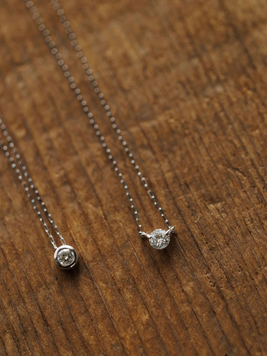 Two delicate Savannah Sevens western life{&}style Petite Diamond Necklaces in 18K white gold with natural round diamond pendants—one bezel set, one prong set—are displayed on a textured wooden surface.