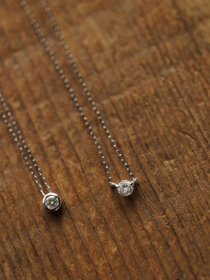 Two delicate Savannah Sevens western life{&}style Petite Diamond Necklaces in 18K white gold with natural round diamond pendants—one bezel set, one prong set—are displayed on a textured wooden surface.
