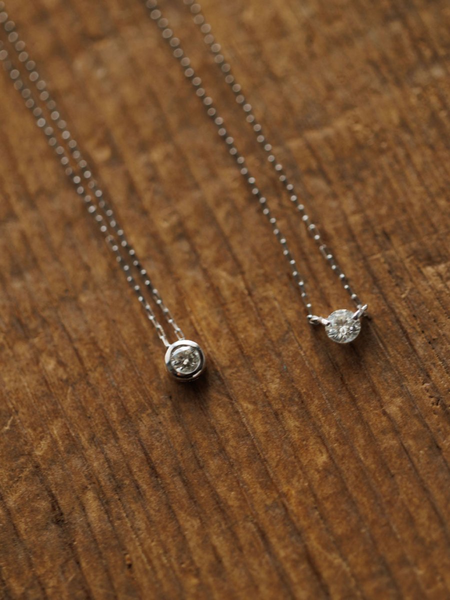 The Petite Diamond Necklace in white gold by Savannah Sevens western life{&}style sparkles delicately, its natural round diamond pendant displayed elegantly on a textured wooden surface.
