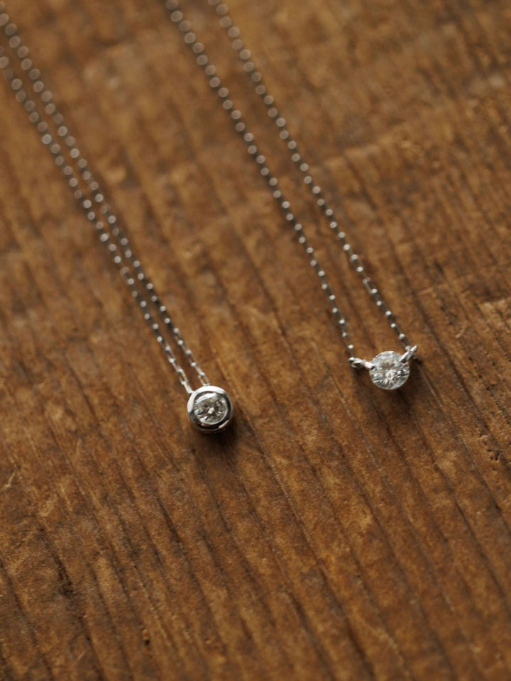 The Petite Diamond Necklace in white gold by Savannah Sevens western life{&}style sparkles delicately, its natural round diamond pendant displayed elegantly on a textured wooden surface.