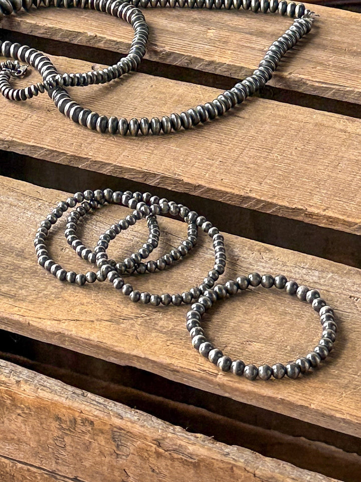 Four metallic beaded bracelets—including the Petros Navajo Pearl Stretch Bracelet by Savannah Sevens western life{&}style and a sterling silver stretch bracelet—are arranged alongside a matching necklace on a wooden surface with wide, evenly spaced planks.