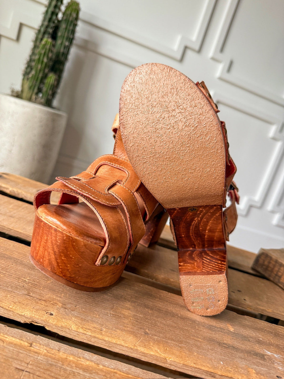A close-up of The Peyette strappy leather heels by Savannah Sevens western life{&}style on wood, highlighting their smooth, textured soles. A potted cactus is visible in the background against a white paneled wall.