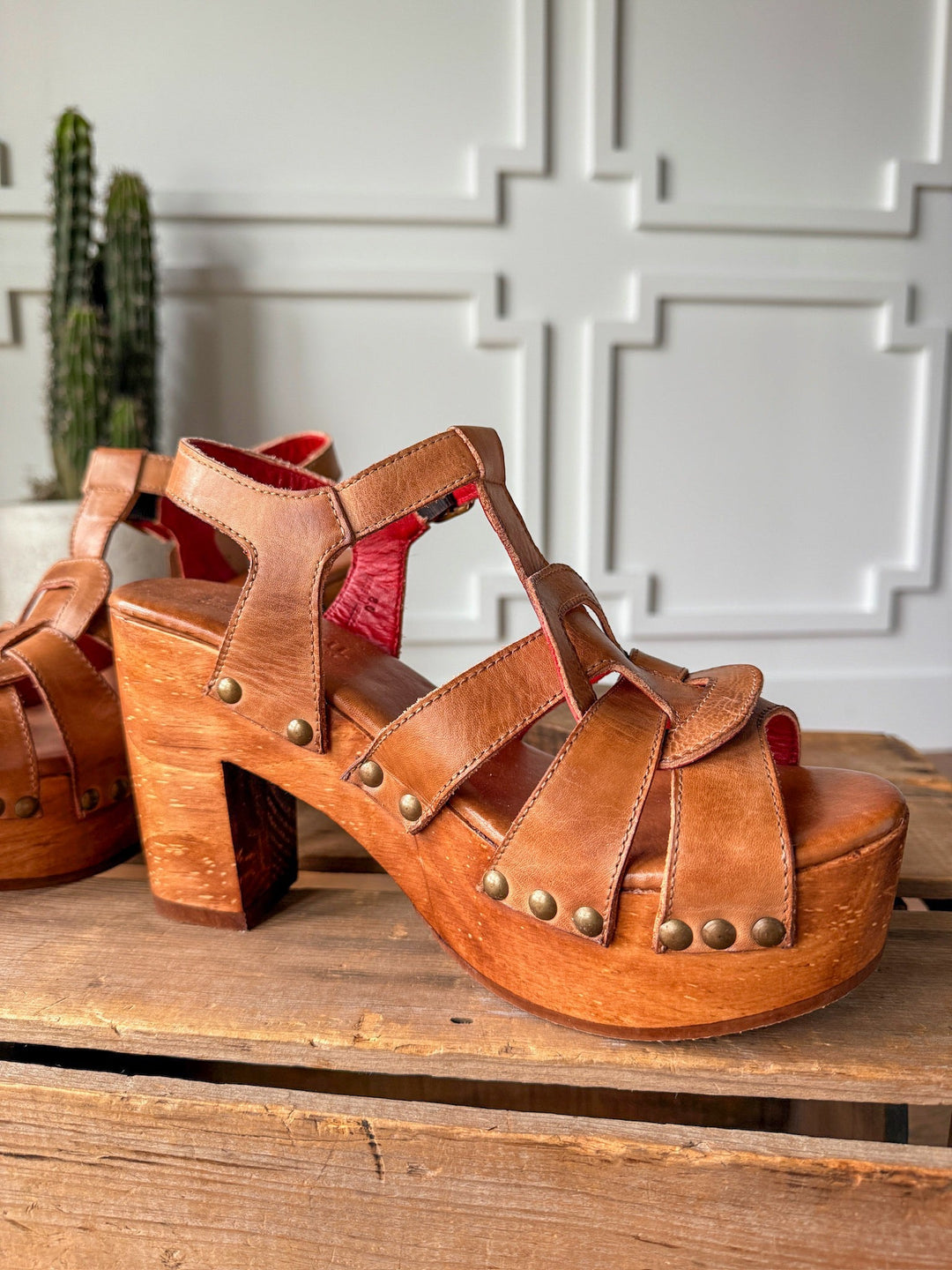 The Peyette by Savannah Sevens western life{&}style, a tan leather sandal with chunky wooden platform heel, metal rivets, and open toe, is shown on rustic wood. Another sandal and a potted cactus appear in the background. Made by BedStu.