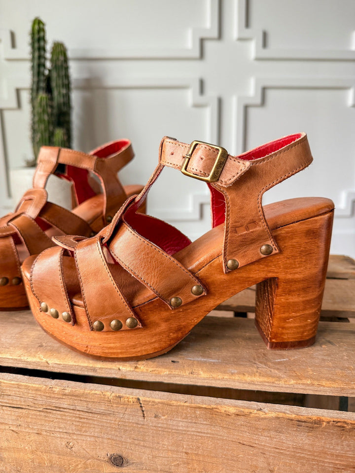 The Peyette by Savannah Sevens western life{&}style: tan strappy leather heels with wooden platforms, metal studs, and buckle straps, shown on a rustic wood surface with a cactus and white paneled wall in the background. Made by BedStu. 