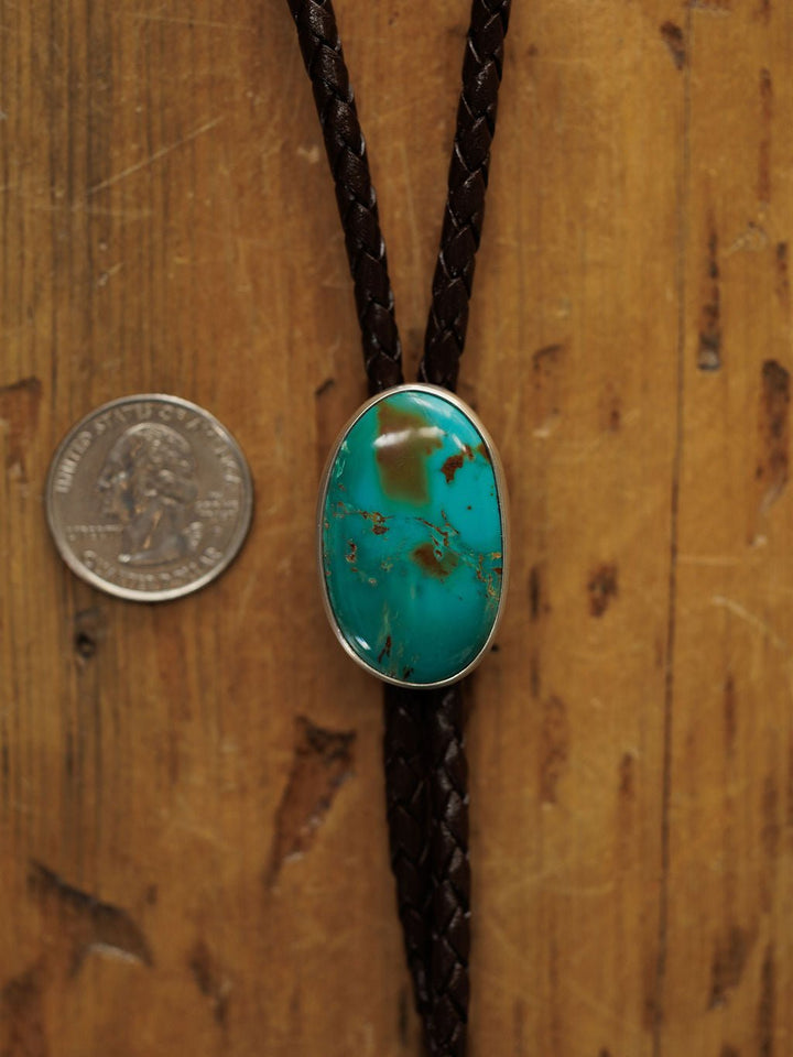 The Peyote Bolo by Savannah Sevens western life{&}style features a handmade leather braid and an oval turquoise stone clasp, shown on wood beside a U.S. quarter for size reference.