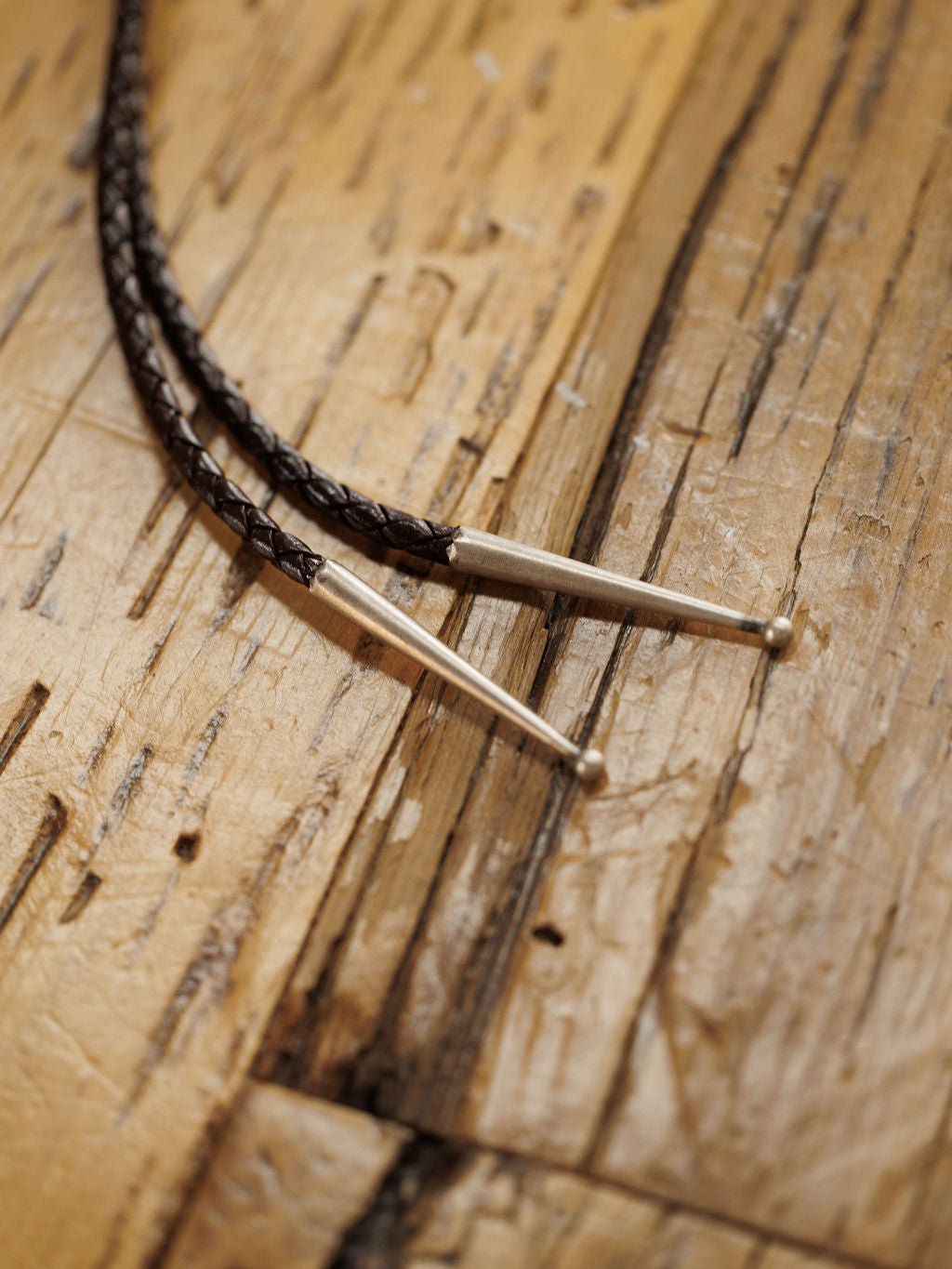 Close-up of two Peyote Bolo braided leather cords with silver metal tips by Savannah Sevens western life{&}style, displayed on a rustic wooden surface with visible grain.
