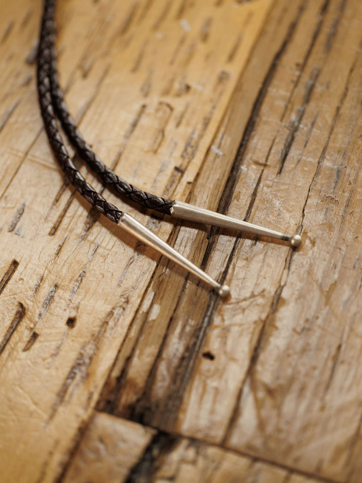 Close-up of two Peyote Bolo braided leather cords with silver metal tips by Savannah Sevens western life{&}style, displayed on a rustic wooden surface with visible grain.