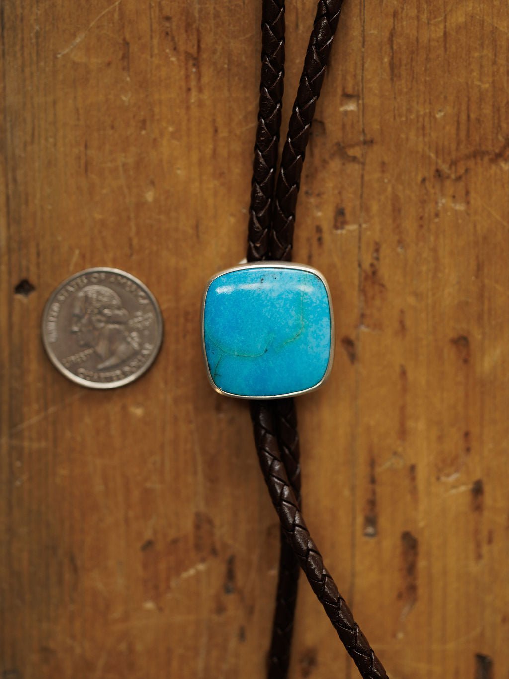 The Peyote Bolo by Savannah Sevens western life{&}style features a turquoise stone in a silver frame on braided dark brown leather, shown beside a U.S. quarter on wood for size reference.