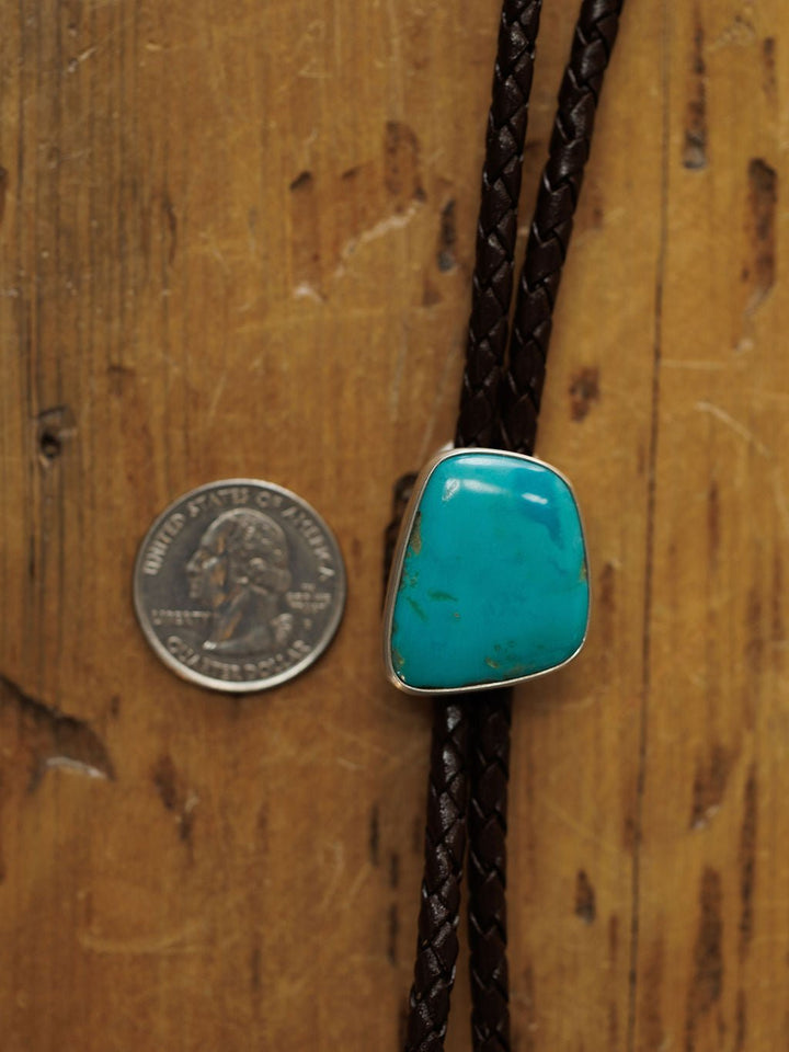 A U.S. quarter is next to the Peyote Bolo by Savannah Sevens western life{&}style, featuring a large turquoise stone set in silver on braided dark brown leather, displayed on a wooden surface.