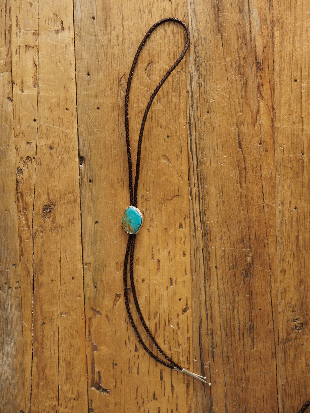 The Peyote Bolo by Savannah Sevens western life{&}style, a handmade leather bolo tie featuring a turquoise stone clasp, is displayed on a rustic wooden surface.