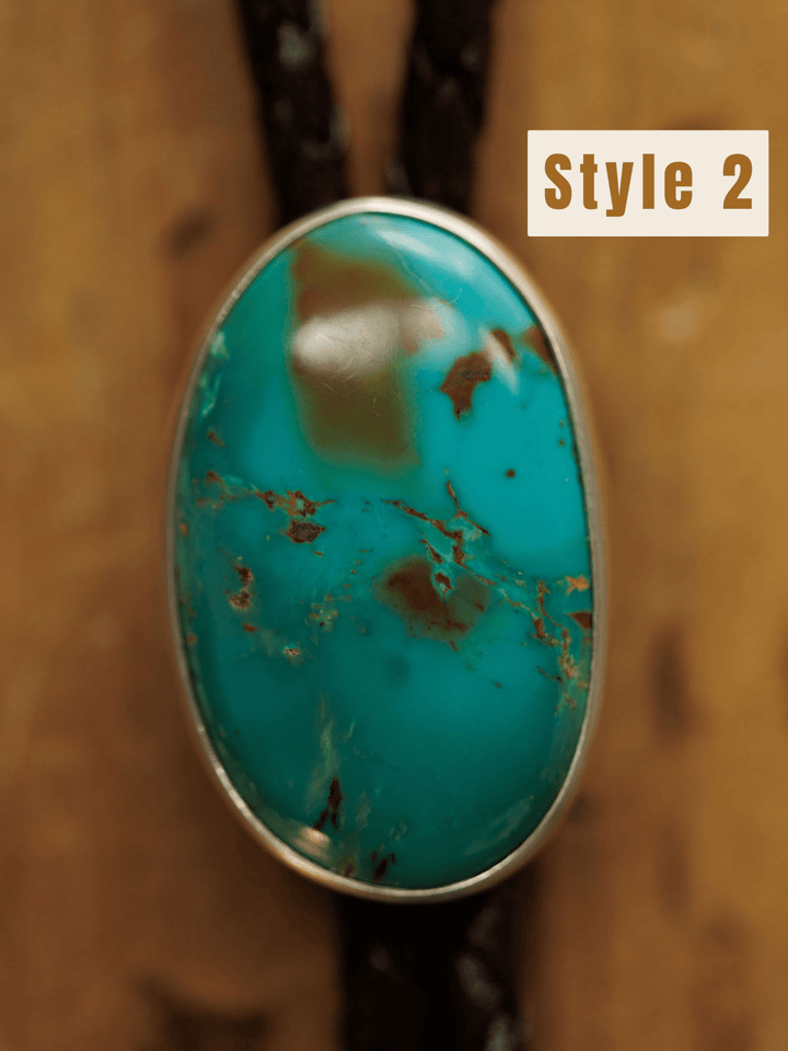 Close-up of the Peyote Bolo by Savannah Sevens western life{&}style: an oval turquoise stone with brown matrix set in a silver bezel on dark braided leather. Blurred wood background; Style 2 label in the top right corner.