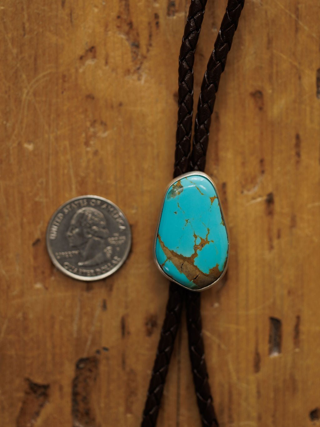 A U.S. quarter sits beside the Peyote Bolo by Savannah Sevens western life{&}style, a handmade bolo tie with a large, irregular turquoise stone on braided dark brown leather, displayed on a wood surface.