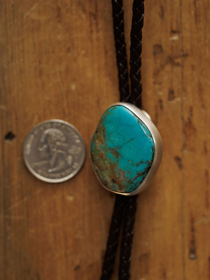 The Peyote Bolo by Savannah Sevens western life{&}style, featuring a large turquoise stone with brown matrix set in silver on braided leather, sits beside a U.S. quarter on wood to show its bold handmade size.