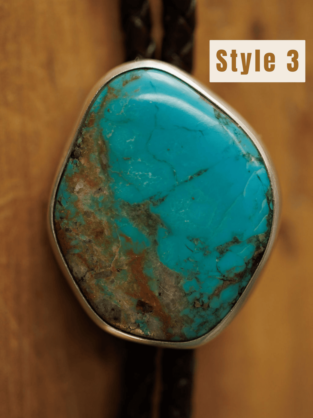 A close-up of the Peyote Bolo by Savannah Sevens western life{&}style, featuring a turquoise stone with brown streaks in a silver bezel on a handmade bolo tie, displayed against wood. "Style 3" appears in a white box at the top right.