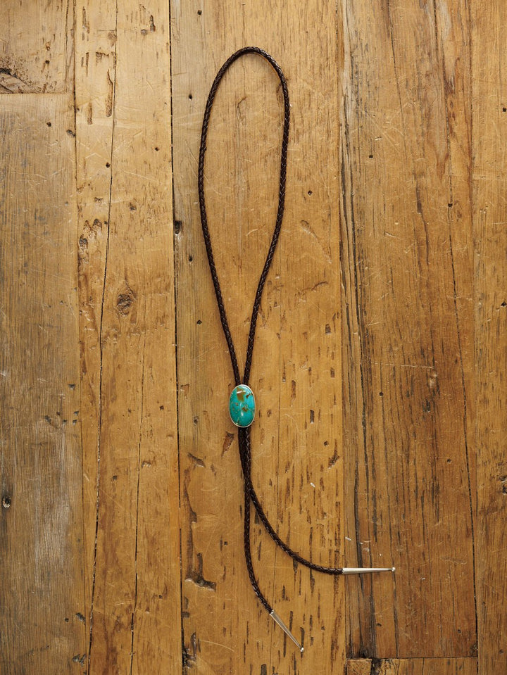 The Peyote Bolo by Savannah Sevens western life{&}style, a handmade leather bolo tie with turquoise stone clasp and metal tips, is displayed on a rustic wooden surface.