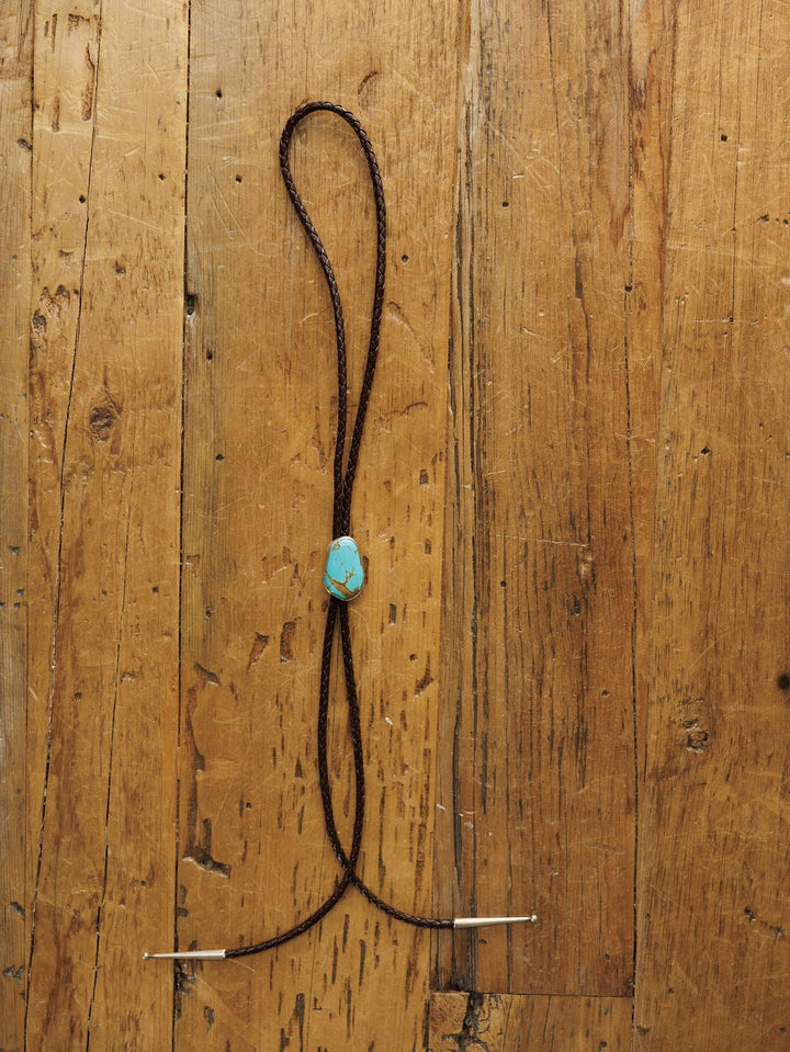 The Peyote Bolo by Savannah Sevens western life{&}style features a handmade leather tie with a turquoise stone clasp, displayed flat on a rustic wooden surface.