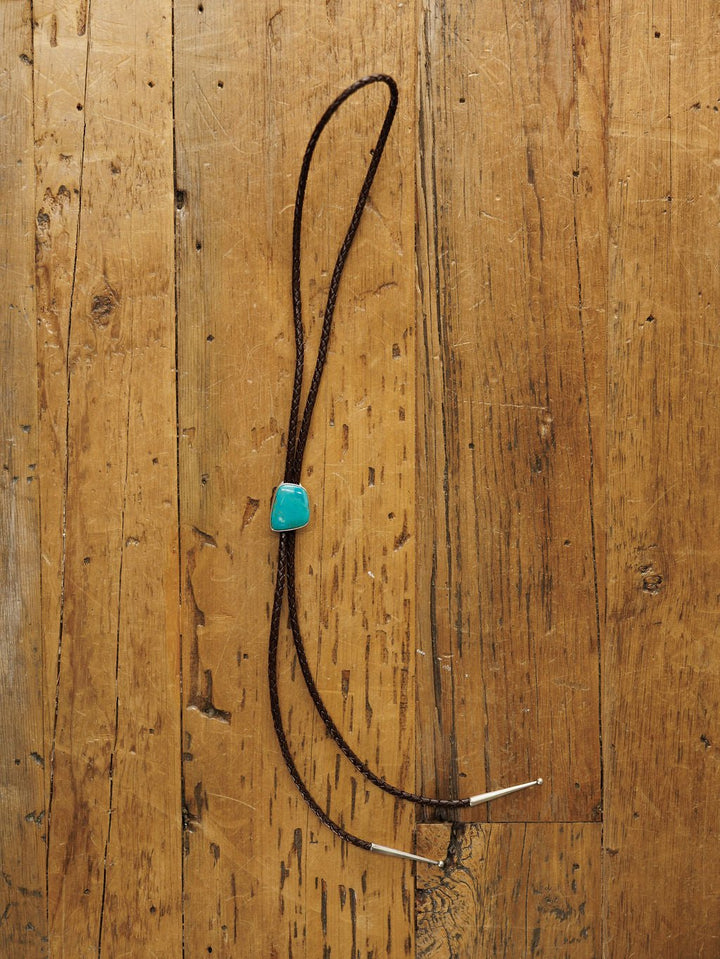 The Peyote Bolo by Savannah Sevens western life{&}style, a handmade leather bolo tie featuring a turquoise stone slide and silver tips, is displayed on a rustic wooden surface.
