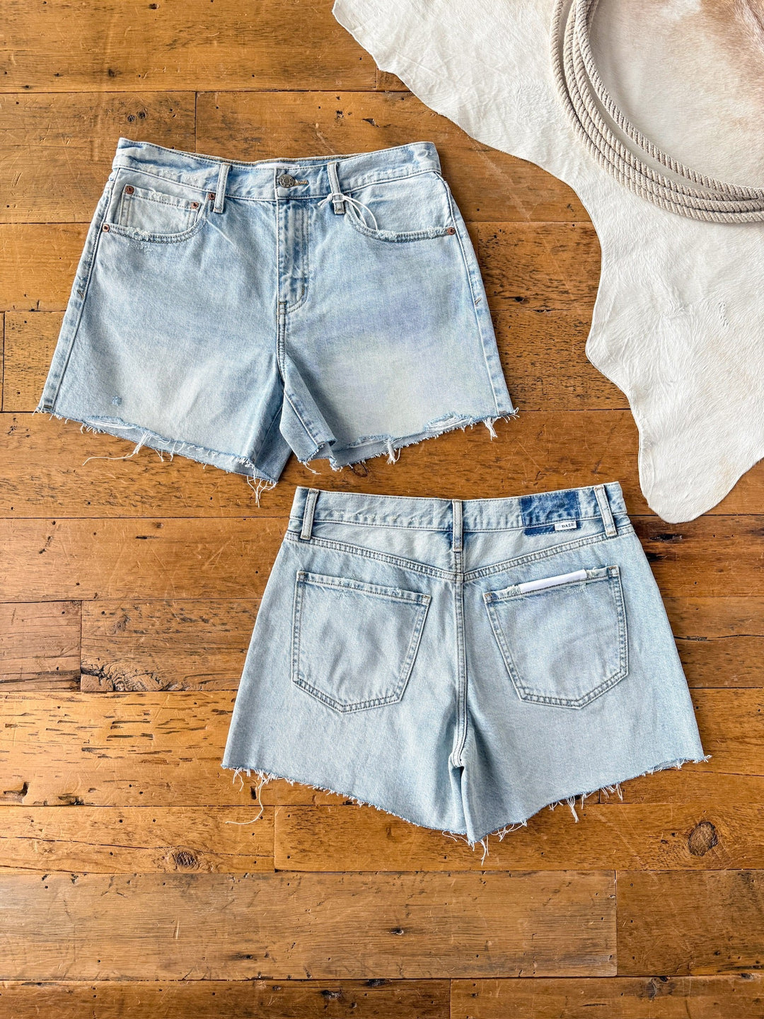 The Peyton Shorts by Savannah Sevens western life{&}style are shown front and back, laid flat on a wooden floor. These are high rise, light wash denim shorts with raw cut hems. A white rug and coiled rope appear in the top right corner.