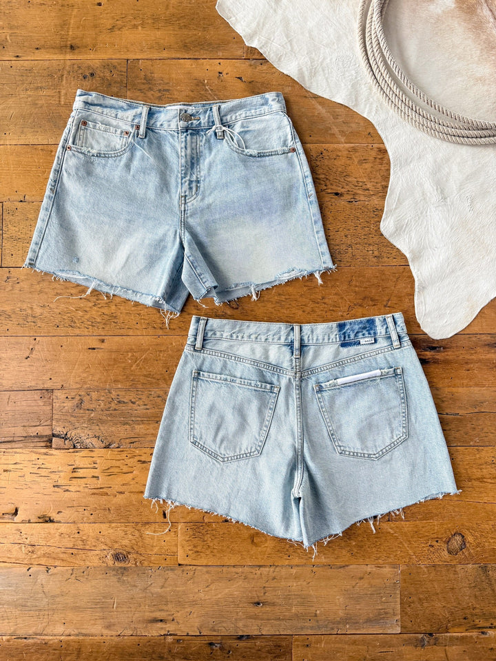 The Peyton Shorts by Savannah Sevens western life{&}style are shown front and back, laid flat on a wooden floor. These are high rise, light wash denim shorts with raw cut hems. A white rug and coiled rope appear in the top right corner.