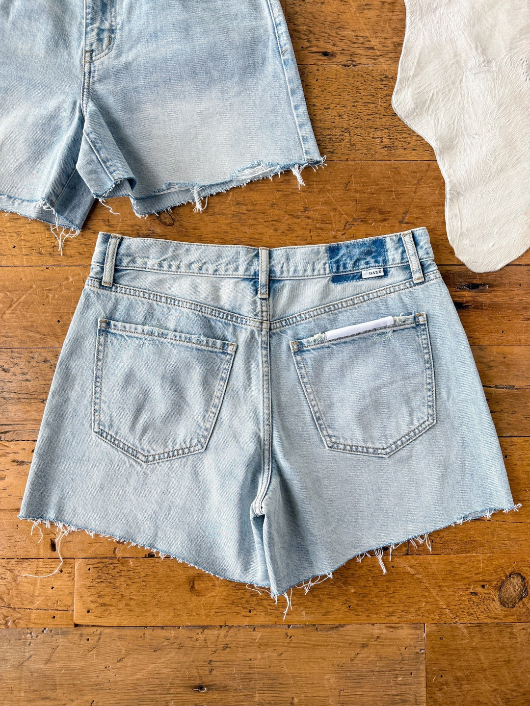 The Peyton Shorts by Savannah Sevens western life{&}style, featuring a light wash and raw cut hem, are laid flat on a wooden surface with parts of another pair of shorts and light-colored fabric visible at the top corners.