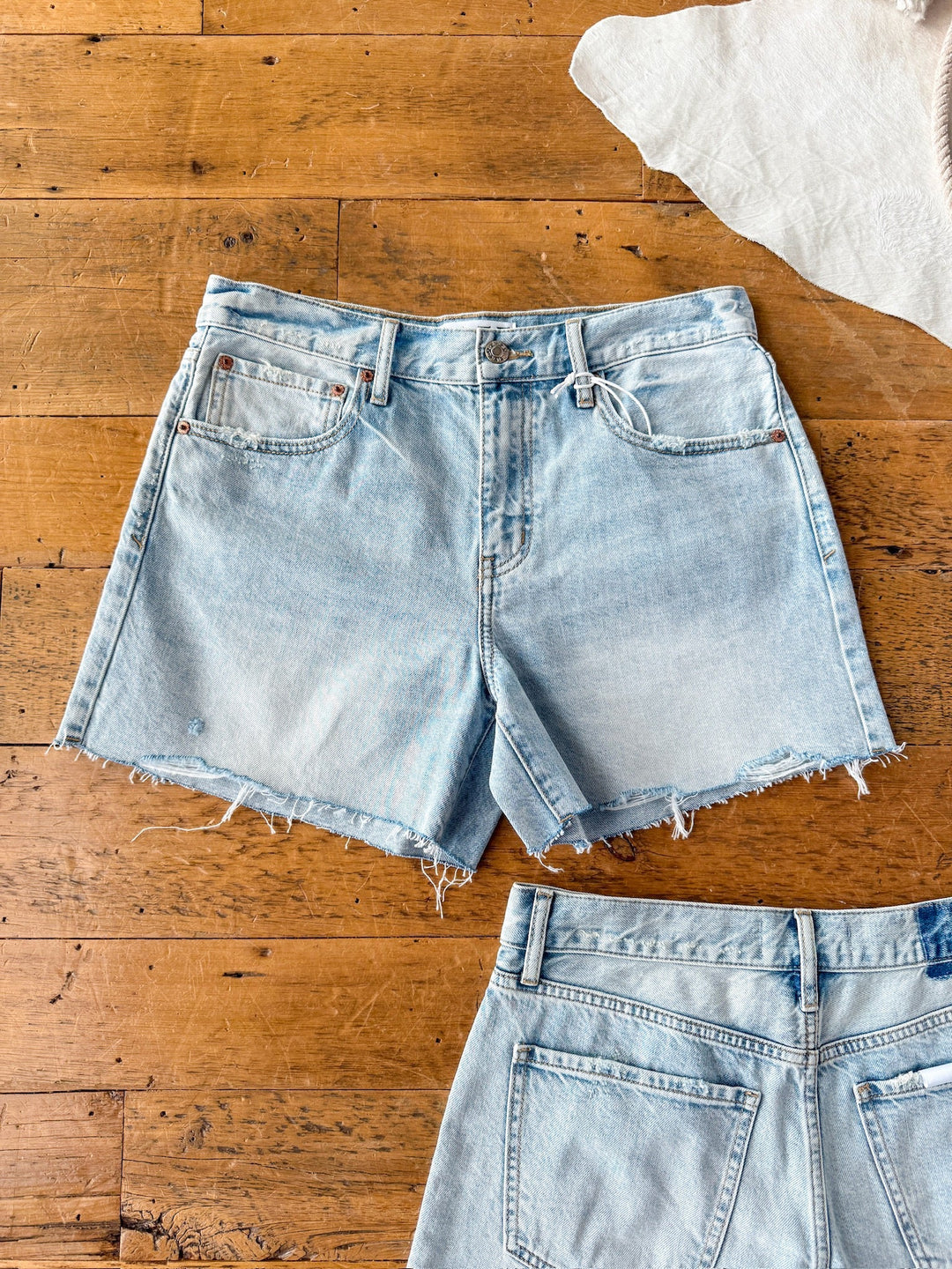 The Peyton Shorts by Savannah Sevens western life{&}style are high-rise denim shorts with a raw cut hem, shown flat on a wooden floor. The full front is visible, with a partial view of another pair’s back at the bottom.
