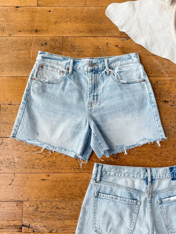 The Peyton Shorts by Savannah Sevens western life{&}style are high-rise denim shorts with a raw cut hem, shown flat on a wooden floor. The full front is visible, with a partial view of another pair’s back at the bottom.