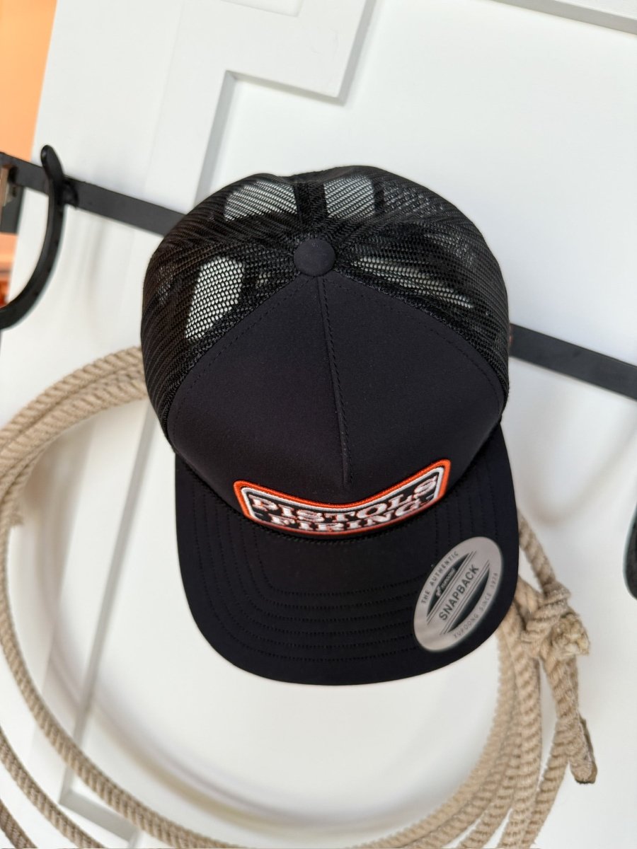 A black Pistols Firing Cap by Savannah Sevens western life{&}style with a red and white patch hangs on a white door above a coiled rope. The cap has a “SNAPBACK” sticker, exemplifying classic Cowboys hat style.