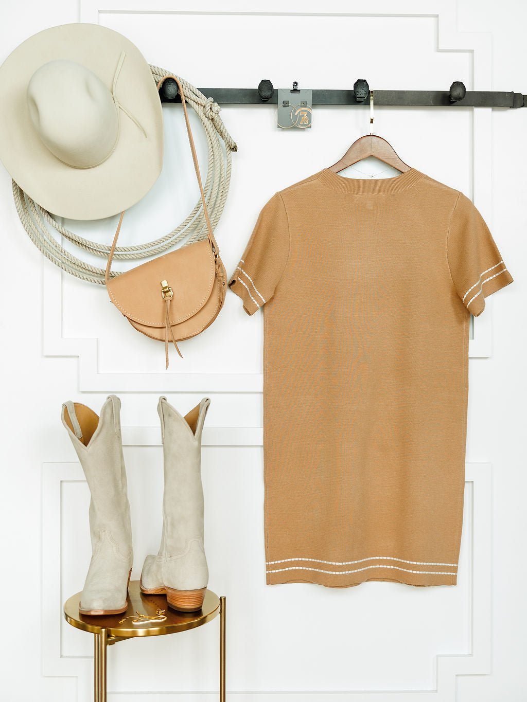 The Platte Tan Stitch Dress by Savannah Sevens western life{&}style hangs on a wall hook next to a beige hat and tan purse, with light beige cowboy boots placed below on a small gold table against a white background.