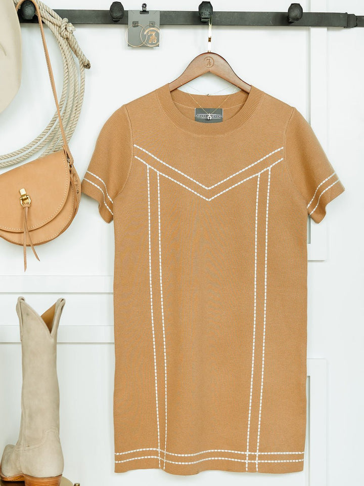 The Platte Tan Stitch Dress by Savannah Sevens western life{&}style hangs on a wooden hanger with a tan purse, beige boots, and a hat nearby, all set against a white wall.