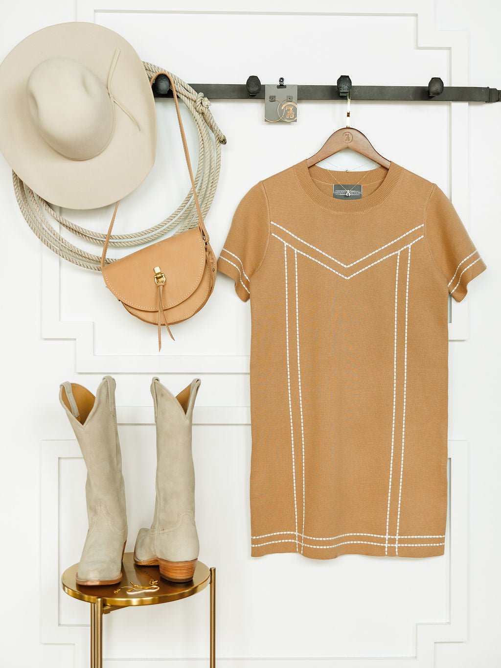 The Platte Tan Stitch Dress by Savannah Sevens western life{&}style hangs on a wall beside a beige hat and tan purse, with light tan cowboy boots placed on a gold stool, all set against a white background.