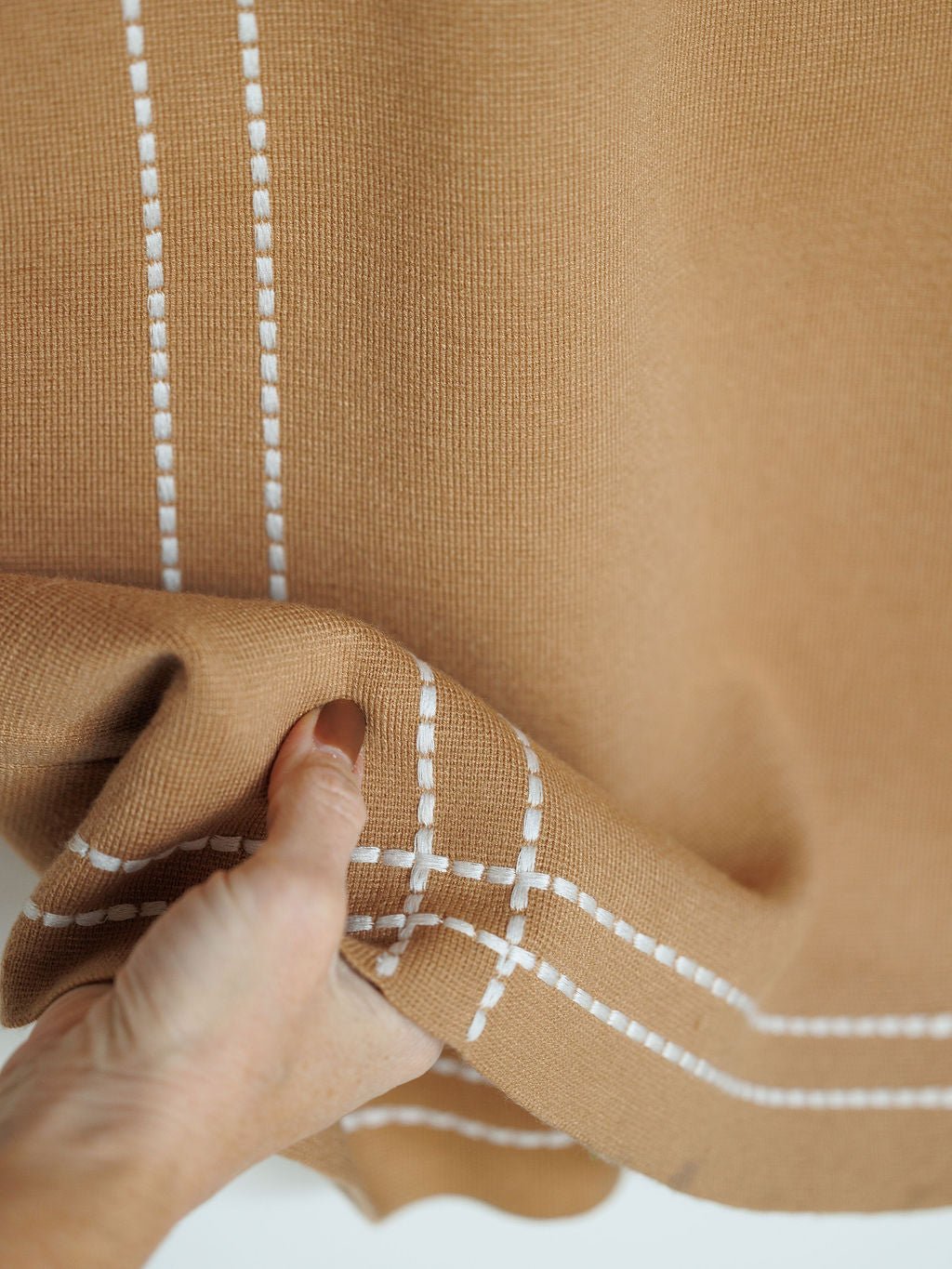 A hand holds the Platte Tan Stitch Dress by Savannah Sevens western life{&}style, featuring tan tri-blend fabric and a white embroidered grid pattern near the edge.