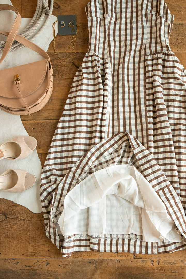 The Plattville Brown Gingham Dress by Savannah Sevens western life{&}style is paired with beige flats, a tan crossbody bag, a rope belt, and earrings, all arranged on a wooden surface.