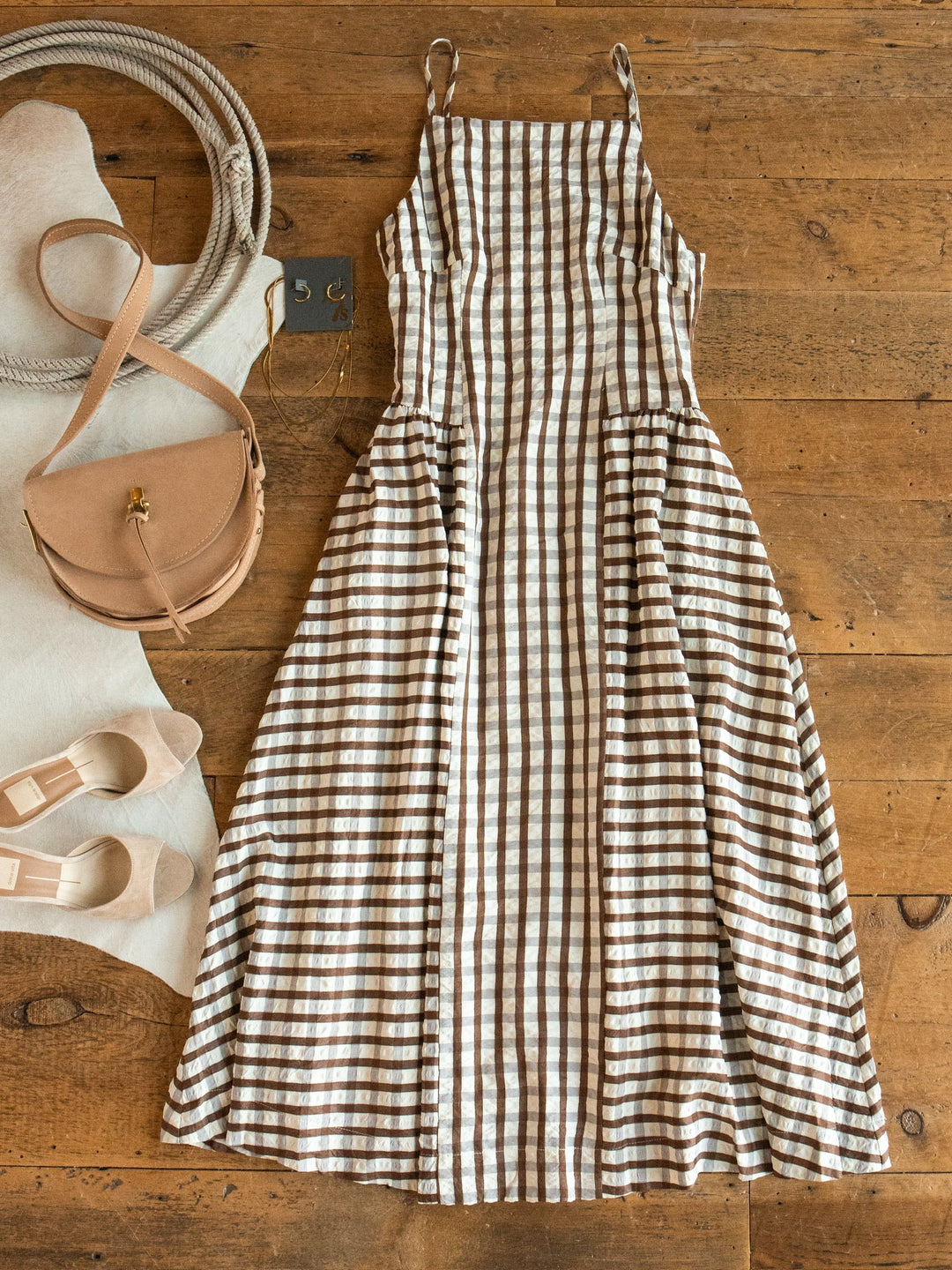 The Plattville Brown Gingham Dress by Savannah Sevens western life{&}style lies flat on a wooden floor, surrounded by beige flats, a tan crossbody bag, a coiled rope, and a white fur rug.