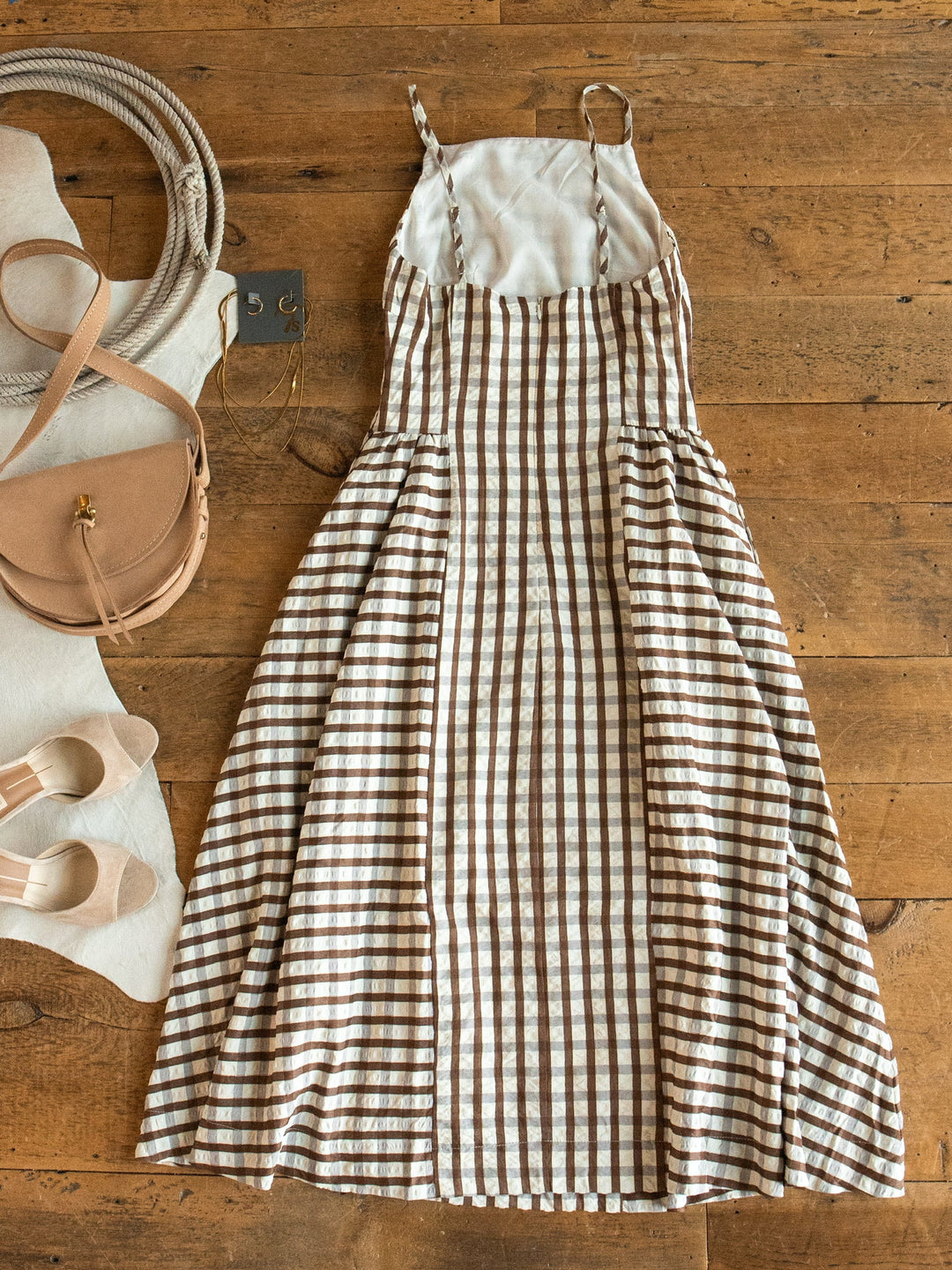 A Plattville Brown Gingham Dress by Savannah Sevens western life{&}style is laid out on a wooden floor, next to beige sandals and a tan crossbody bag.