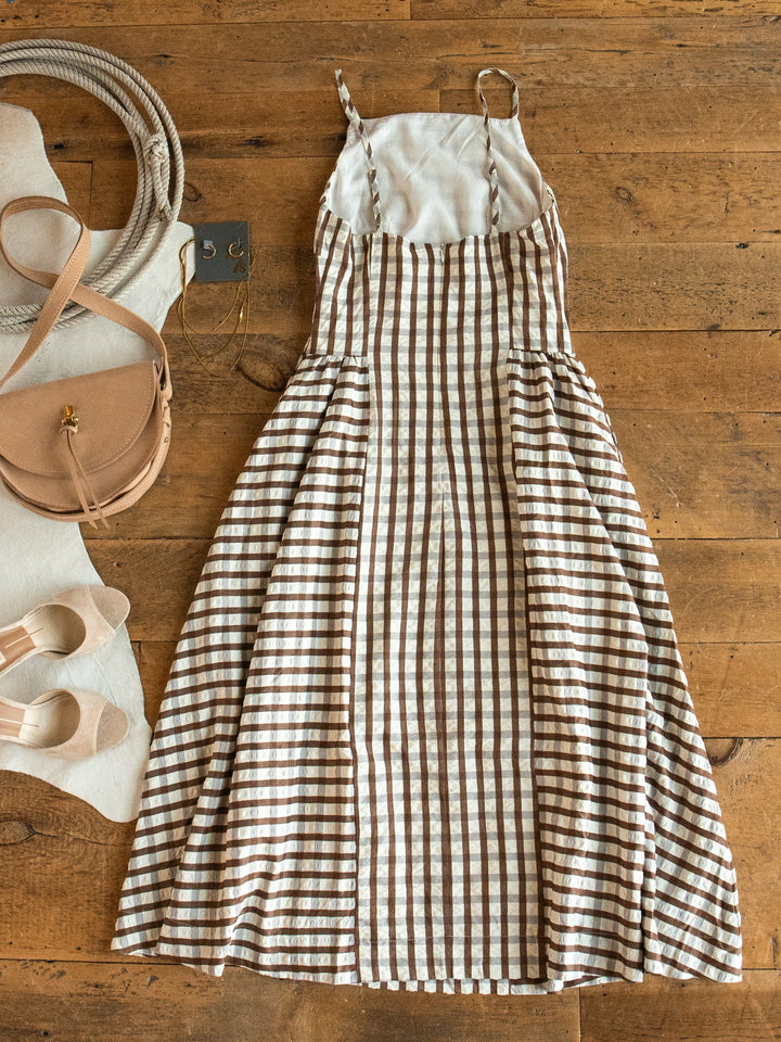 A Plattville Brown Gingham Dress by Savannah Sevens western life{&}style is laid out on a wooden floor, next to beige sandals and a tan crossbody bag.