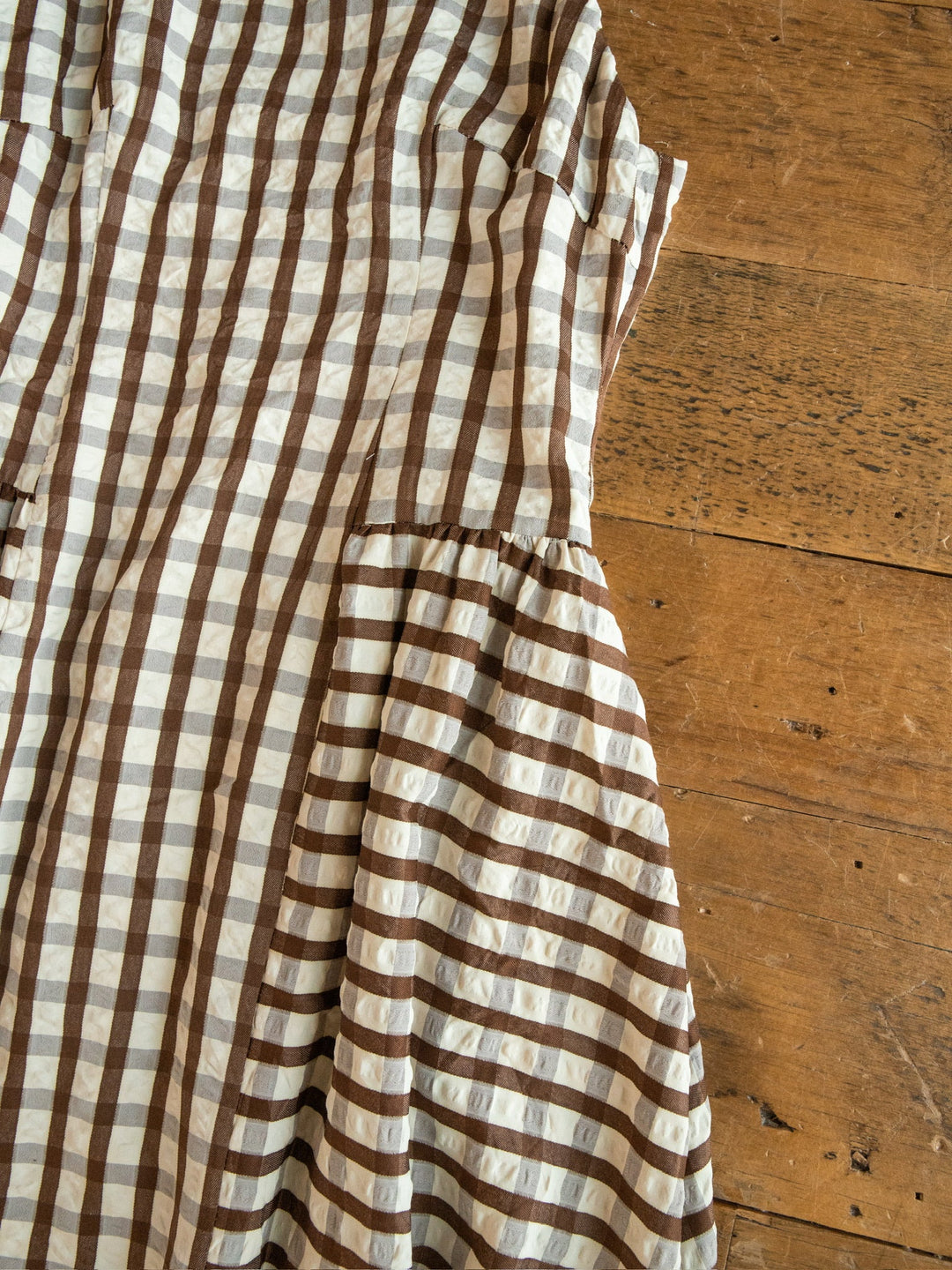 A partially visible Plattville Brown Gingham Dress by Savannah Sevens western life{&}style, made from a cotton blend, lies on a wooden floor.