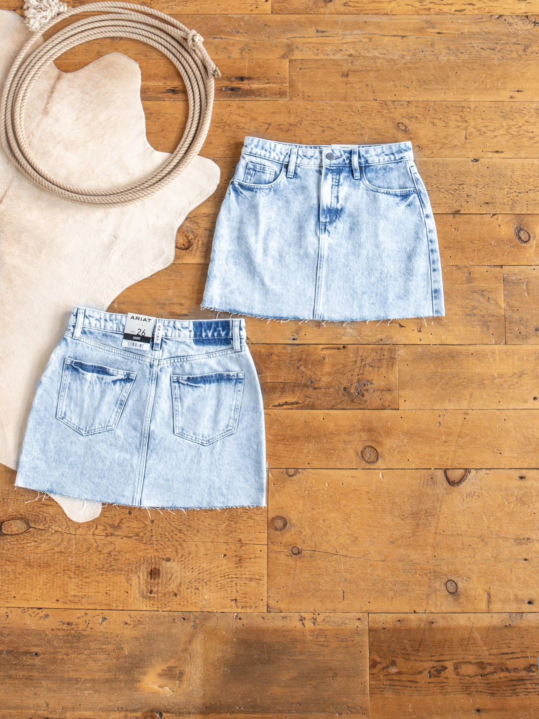 Two Polson Skirts by Savannah Sevens western life{&}style, in light blue acid wash denim with raw cut hems, are displayed front and back on a wooden floor beside a coiled rope and a light animal hide rug.
