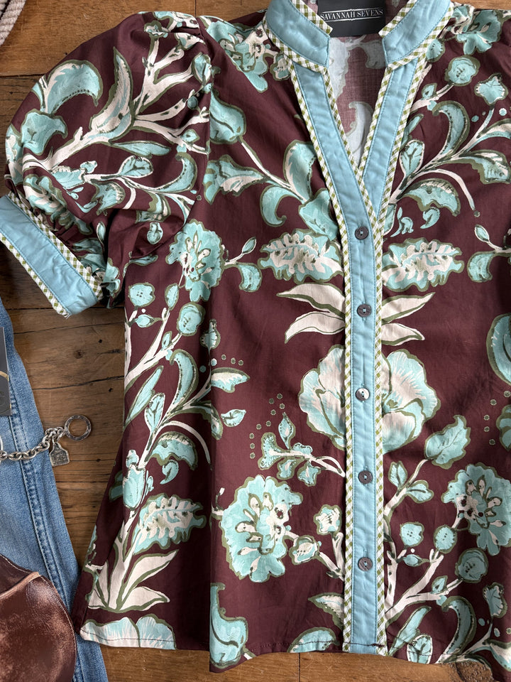 The Pomaria Floral Puff Sleeve Top from Savannah Sevens western life{&}style, featuring a maroon background with a light blue floral pattern and blue trim along the collar, sleeves, and button placket, is displayed on a wooden surface beside denim jeans and a brown leather bag.