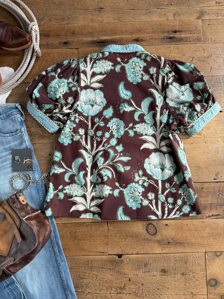 The Pomaria Floral Puff Sleeve Top by Savannah Sevens western life{&}style, featuring a brown base with light blue floral patterns, is displayed on a wooden floor beside blue jeans, a brown leather bag, a belt, and part of a shoe.