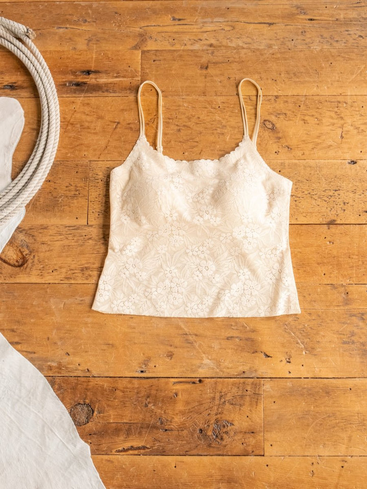 The Pomona Bralette by Savannah Sevens western life{&}style, featuring a cream lace pattern and adjustable straps, is laid flat on a rustic wooden floor with a coiled rope and white fabric partially in view.