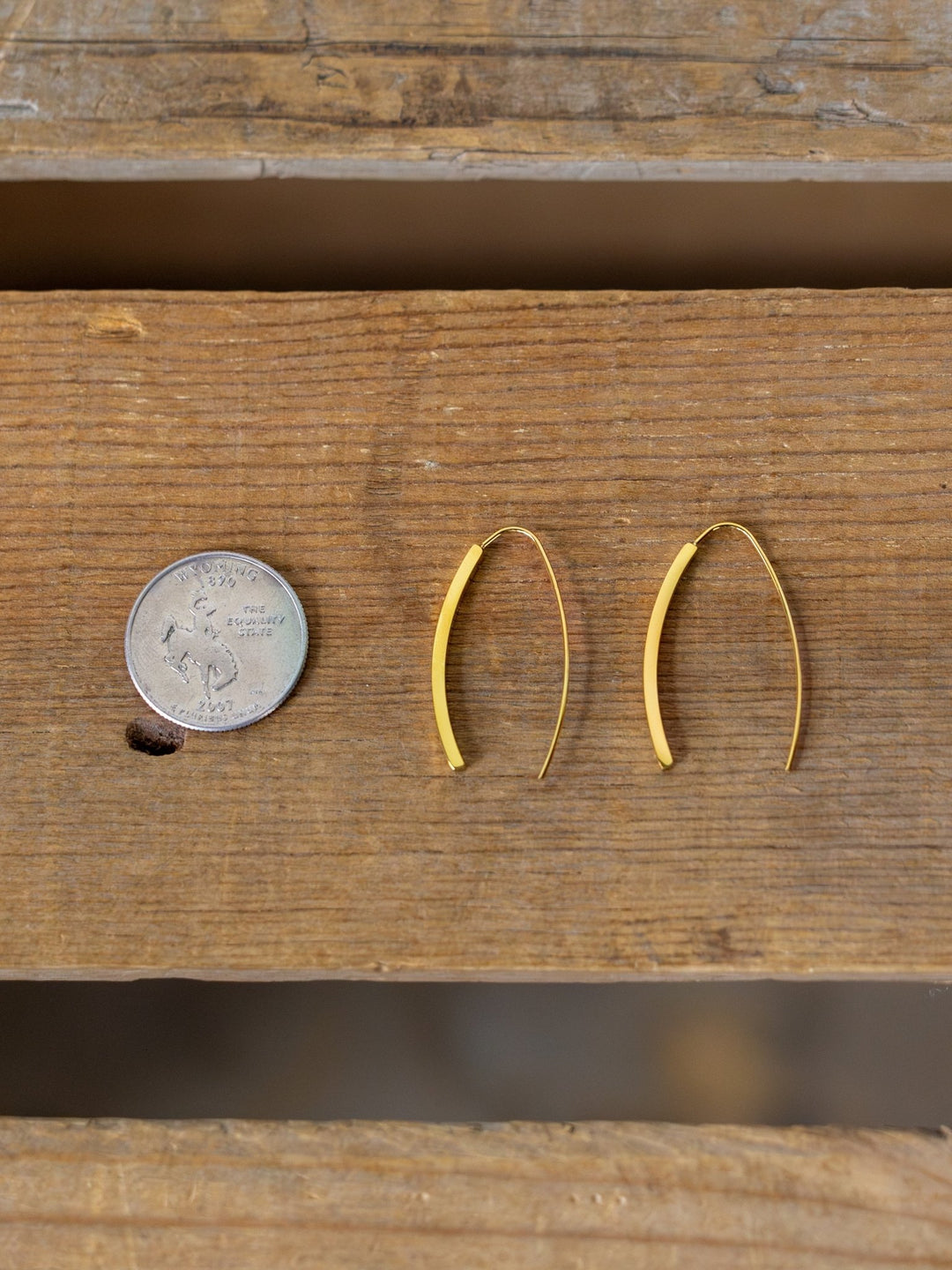 A U.S. quarter sits beside Pondera Gold Earrings from Savannah Sevens western life{&}style on wood, highlighting their size. These earrings are waterproof, hypoallergenic, and designed for stylish comfort.