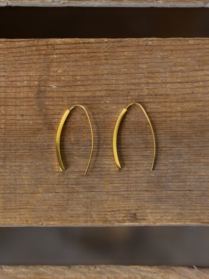 The Pondera Gold Earrings by Savannah Sevens western life{&}style are minimalist, curved hypoallergenic drops shown on a rustic wooden surface.